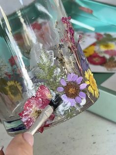 a person holding a glass with flowers on it and paintbrush in the bottom half