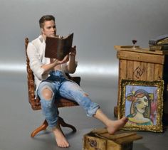 a man sitting in a chair reading a book next to a miniature desk with a painting on it