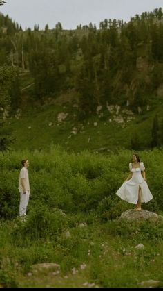 two women in white dresses are walking through the grass near some rocks and trees on a hill