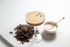 a drink in a glass next to some coffee beans on a white surface with a spoon