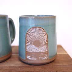 two coffee mugs sitting next to each other on top of a wooden table with a white wall in the background