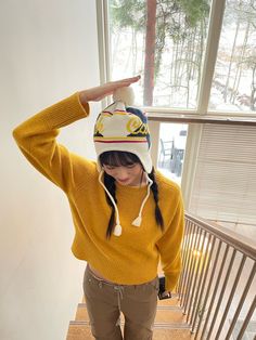 a woman in a yellow sweater and hat is standing on the stairs with her hands behind her head