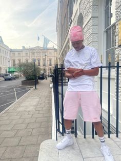 a man in pink shorts and a white shirt is standing on the sidewalk looking at his cell phone