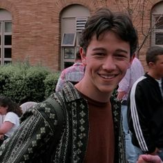 a young man standing in front of a brick building smiling at the camera with other people behind him