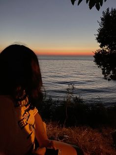 a woman sitting on top of a grass covered hillside next to the ocean at sunset