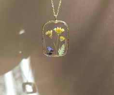 The necklace was made with handpicked and pressed yellow dandelion and epoxy resin. The flower is encased in gold color rectangle frame with clear resin. The resin protects the flower and at the same time it magnifies its beauty.  The sizes of the pendant is 3 cm x 4.5 cm. The chain length is 41 cm with 4 cm extension part and it's closed with a lobster clasp. (Totally 45 cm.) This dainty botanical jewelry can be: - A perfect dandelion gift for flower lovers, - a floral birthday gift, an anniversary gift, a mother's day gift or a christmas gift for her; for your girlfriends, sister, daughter or mom, - or a gift for yourself to wear a bit of nature on you and feel the spring always with you.   Since every flower is unique, every piece in my shop is just one in stock. If you want it to be yo Dainty Pressed Flowers Pendant Jewelry, Delicate Pressed Flower Pendant Jewelry, Yellow Pressed Flowers Jewelry, Green Pressed Flowers Round Pendant Jewelry, Dandelion Necklace, Nature-inspired Flower Pendant Necklace With Pressed Flowers, Botanical Jewelry, Clear Resin, Little Flowers