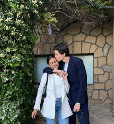 a man and woman standing next to each other in front of a stone wall with flowers on it