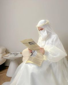 a woman in a white dress and veil is reading a book while sitting on a chair