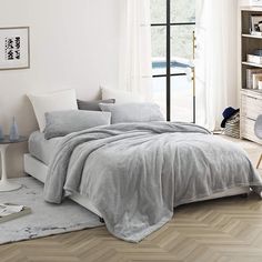 a bed in a bedroom with grey linens and pillows on the floor next to a window