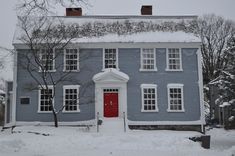 a blue house with white trim and red door is covered in snow as it sits next to a tree