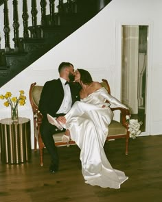 a bride and groom sitting on a chair in front of a stair case, kissing