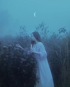 a woman in white dress standing next to bushes and flowers with the moon behind her