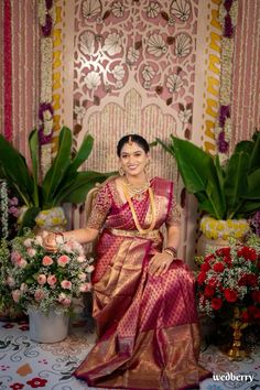 a woman in a pink and gold sari sitting on a chair next to flowers