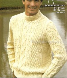 a man wearing a white sweater and hat in front of a body of water with grass