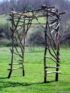 an outdoor sculpture made out of branches in the middle of a green field with trees behind it