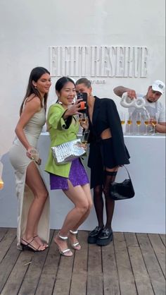 three women standing next to each other with drinks in their hands and one woman taking a selfie