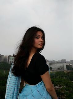 a woman with long dark hair wearing a black top and blue skirt standing in front of a cityscape