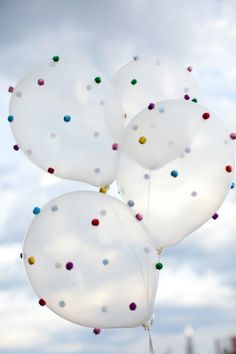 three white balloons with multicolored confetti on them in the air against a cloudy sky