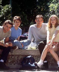 four people sitting on a stone bench in front of some trees and bushes, with one person sprinkled with water