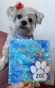 a small white dog holding up a piece of art