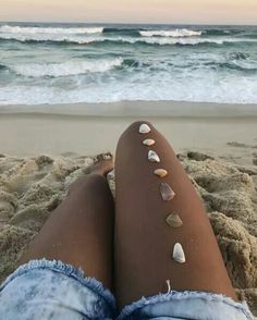 a woman's legs with seashells on them sitting in the sand at the beach