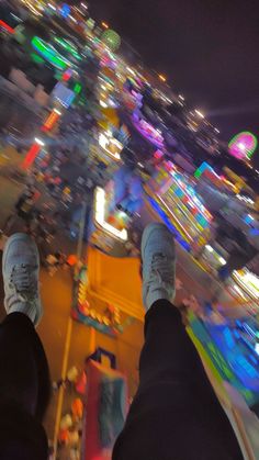 the feet of a person standing on top of a carnival ride at night with brightly lit rides in the background