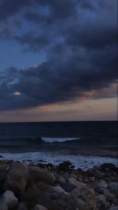 the sky is dark and cloudy over the ocean with rocks on the beach in front of it