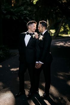 two men in tuxedos standing next to each other on a road with trees behind them