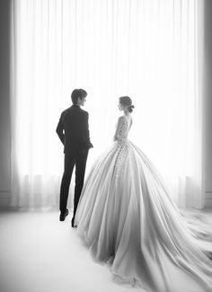 a man standing next to a woman in a wedding dress near a window with curtains