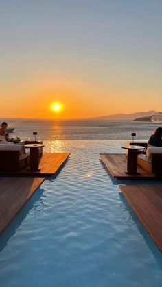 two people sitting on lounge chairs next to a swimming pool at sunset with the sun setting