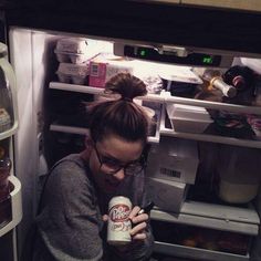 a woman drinking from a can in front of an open refrigerator