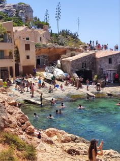 many people are swimming in the water near some buildings and cliffs, while one person is taking a photo with his cell phone