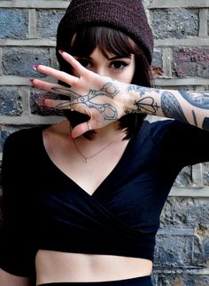 a woman with tattoos on her arm and hand covering her face while standing in front of a brick wall