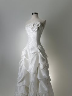 a white wedding dress with ruffles and flowers on the bustle, is displayed in front of a gray background