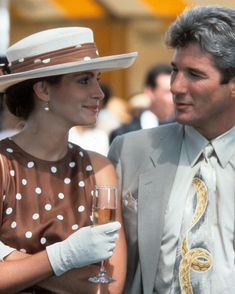 a man standing next to a woman wearing a hat and holding a glass of wine