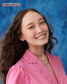 a woman with long brown hair wearing a pink shirt and pearl necklace smiling at the camera