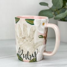 a coffee mug with the word ruby painted on it next to a potted plant