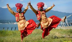 two men dressed in red and gold dance on grass with mountains in the background at an outdoor event