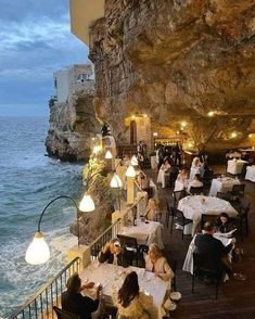 people sitting at tables near the ocean in front of an cliff side restaurant with lights on