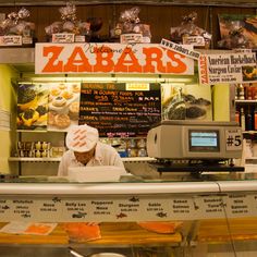 a man working behind the counter at a food stand in a market, with an orange sign above it that says zabars