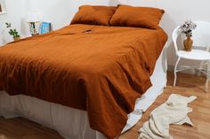 a bed with an orange comforter in a white room next to a chair and potted plant