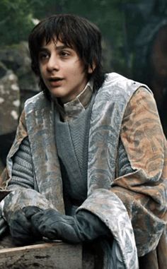 a young man dressed in medieval clothing sitting on a stone bench with his arms crossed