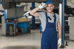 a man in overalls and gloves standing under a car with his hands on his head