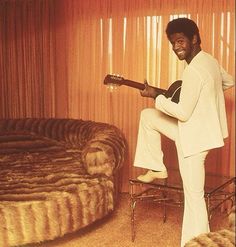 a man in white is playing an acoustic guitar and sitting on a bed with his legs crossed