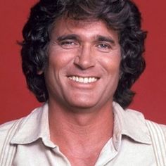 a man with long hair smiling at the camera in front of a red background wearing a white shirt