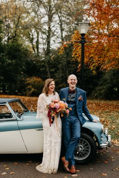 a bride and groom standing next to an old car in the fall with leaves on the ground