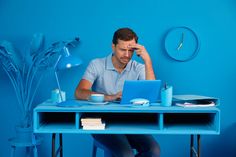 a man sitting at a blue desk with a laptop in front of him and a clock on the wall behind him