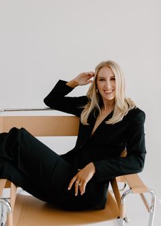 a woman sitting on top of a wooden chair in front of a white wall with her legs crossed