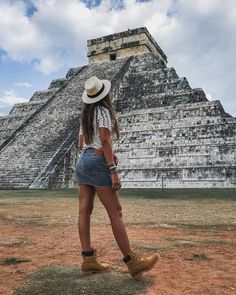 a woman walking in front of an ancient pyramid with a hat on her head and boots