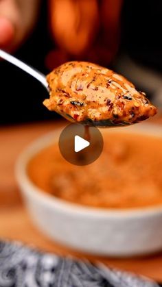 a person holding a spoon full of soup with chicken on it's side and another bowl in the background